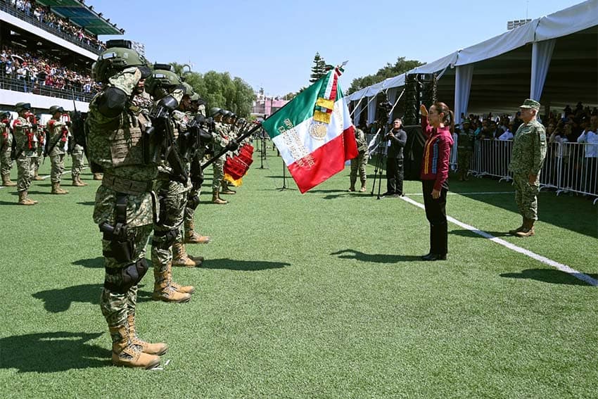 Mexico's President Sheinbaum returning the salute of Mexican marines in unform line in a row, with one holding the Mexican flag in a stadium in Queretaro