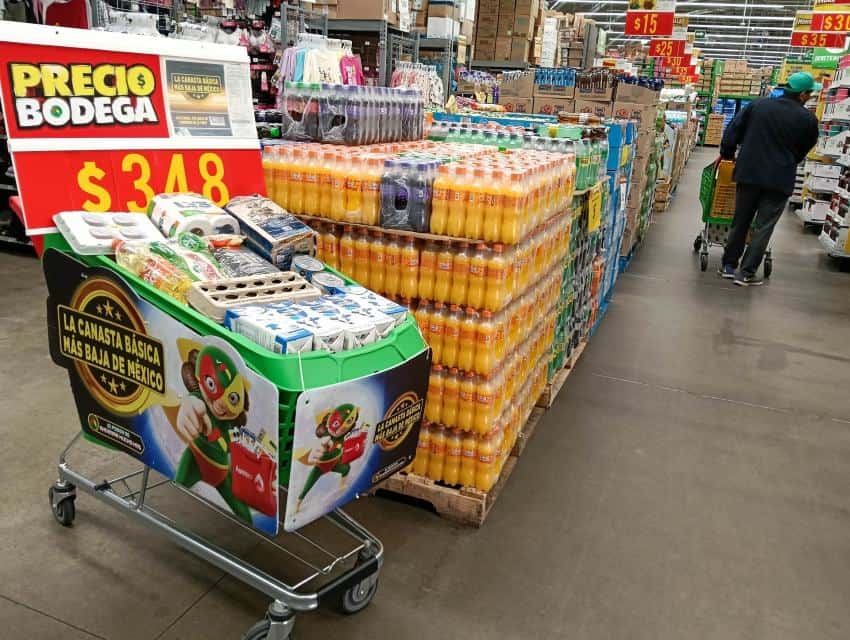 Grocery cart filled with items from the Mexican food basket inside a Mexican supermarket with aisles of grocery items on display. A single shopper pushing a shopping cart is in the background.