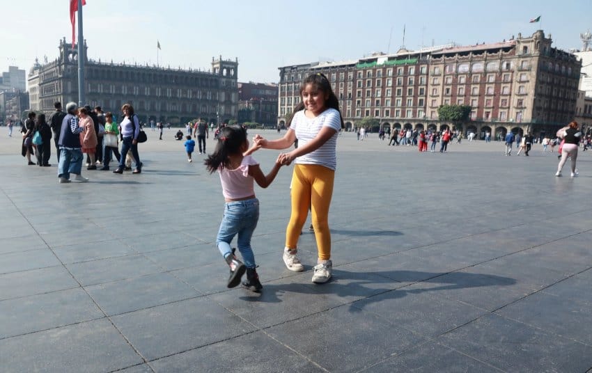 Two young Mexican girls, one around 10 years old and one around five years old, caught by the camera in the middle of dancing together while holding hands in the middle of Mexico City's expansive pedestrian square, The Zocalo. In the background, small groups of people are hanging out in the square. and behind them are large multistory buildings from the colonial era of Mexico.