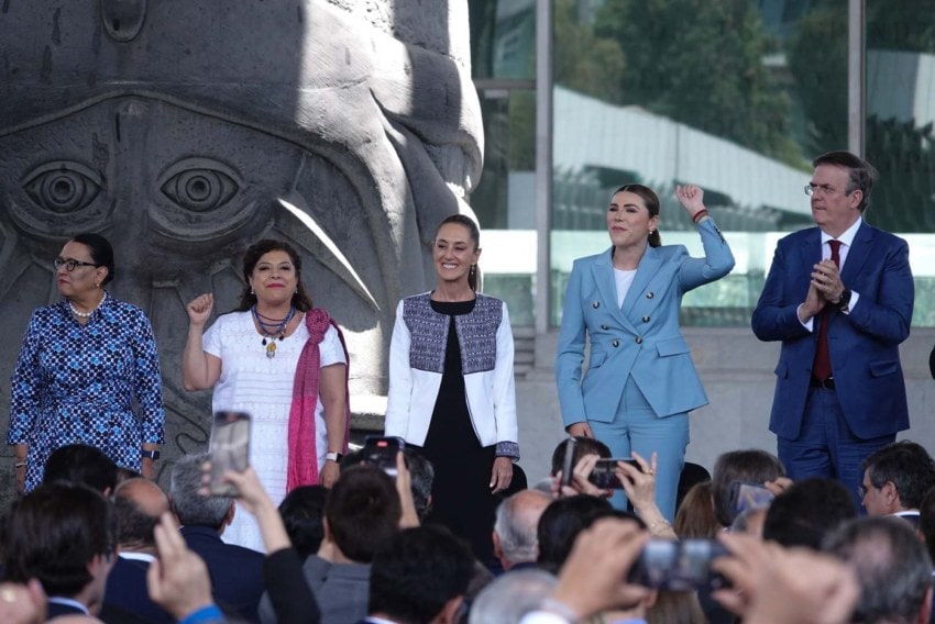 Mexico's President Claudia Sheinbaum Plan Mexico event with her at the center of a line of Mexican officials onstage. Two of them are holding up their closed fists as if to cheery, and one is applauding.