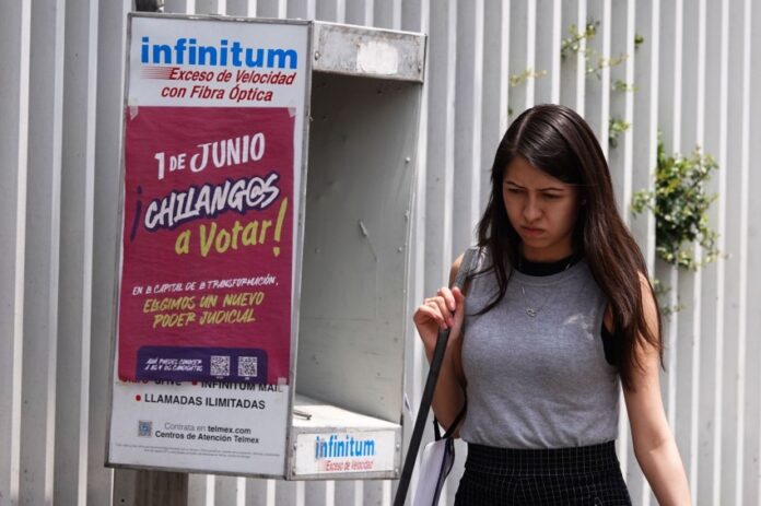 A Young woman in a gray sleeveless top and a black skirt and carrying a white messenger bag over her shoulder walks past a telephone booth in Mexico City that has a sign urging Mexico City residents to vote in the upcoming judicial election on June 1, 2025.