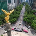 An aerial view of the Angel of Independence monument on Reforma Avenue in Mexico City, looking down the tree-lined boulevard surrounded by tall city buildings.