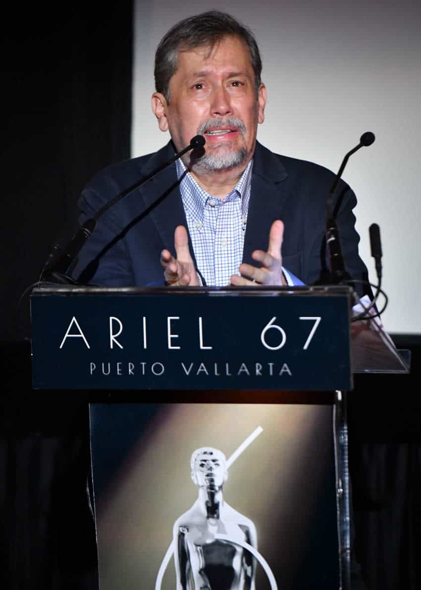 Armando Casas, president of the Mexican Academy of Arts and Cinematographic Sciences, speaking at a podium for film award nominations for the 67th annual Ariel film awards of Mexico. The podium has an image of the Ariel award, which looks a bit like an Oscar trophy but in silver, and a sign that says "Ariel 67 Puerto Vallarta"