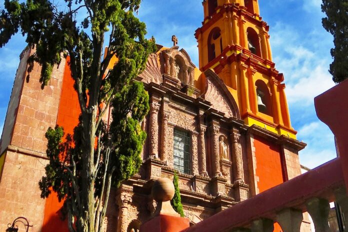 Close-up of a historic church in San Miguel de Allende, showcasing its distinctive orange and yellow colors, ornate facade, and bell tower, with a cypress tree in the foreground.