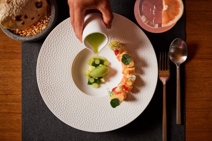 A fine dining presentation of a dish involving pieces of sushi and fresh fruit on a white plate on a placemat next to a cocktail and tortillas on an artisan stone bowl.