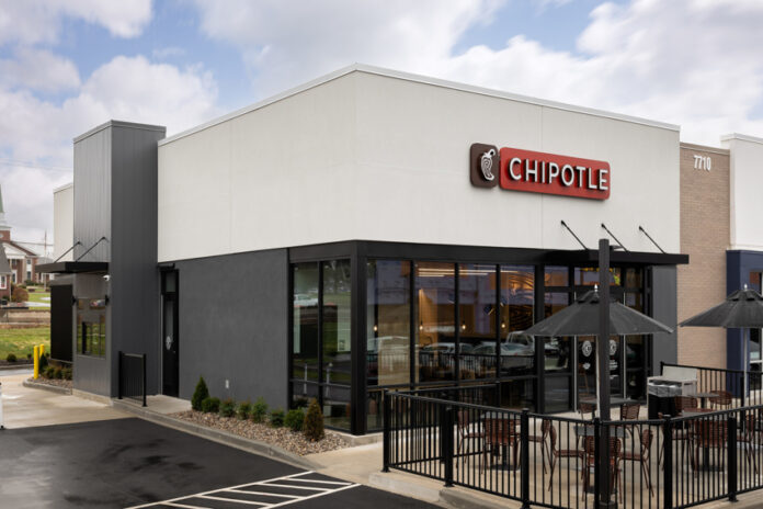 A concrete black and white Chiptole restaurant front, with the red Chipotle logo with white letters sitting on the building's top center. Outside, next to the front door is a small patio with tables for outdoor dining.