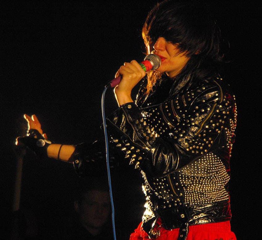 Karen O, lead singer of the Yeah Yeah Yeahs band, a woman with dark, shaggy hair. She is singing into a microphone, illuminated by a spotlight against a dark background. She is wearing a heavily studded black leather jacket over a red garment.