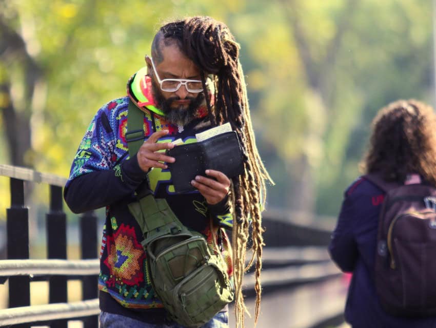 A young Mexican man in a colorful sweater and white reading glasses and who sports dreadlocks down to his waist looks over the contents of his wallet on campus outside the National Autonomous University of Mexico's campus center. Nearby, a young female student wearing a jacket and backpack walks in the other direction