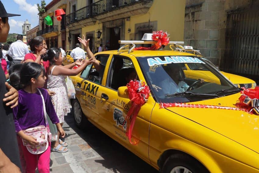 Pedestrians wave to decorated taxi in Oaxaca, cab driver throws gifts from window