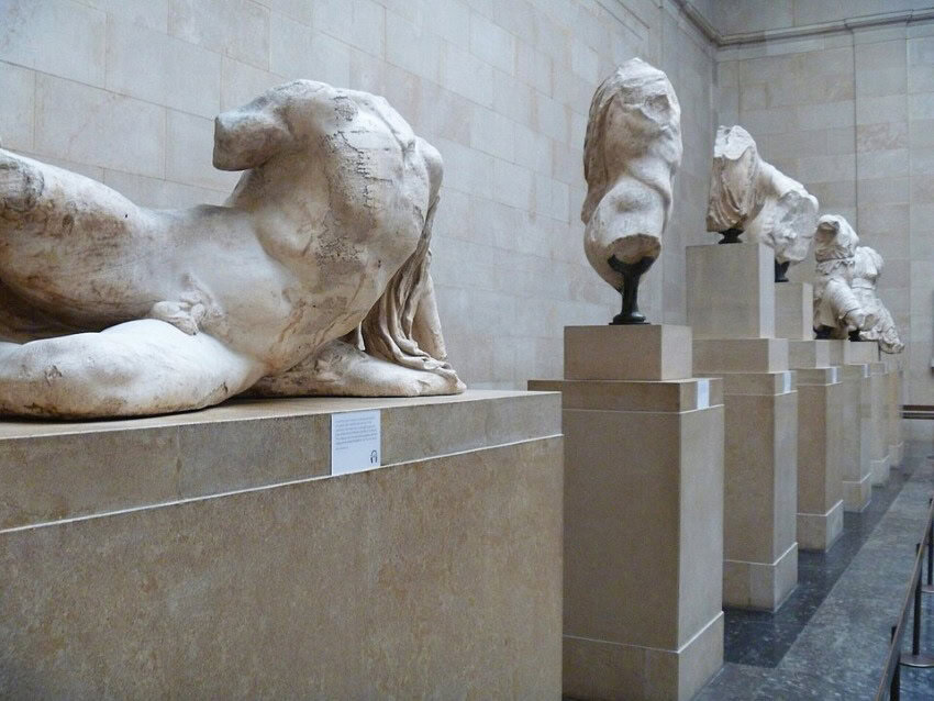 Ancient Greek busts and torsos missing heads, arms, torsos, on display in a row on individual pedestals.