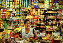 My family runs a convenience store in México state: Here’s everything I’ve learned A Mexican middle-aged woman with a warm smile stands behind a counter laden with colorful packaged goods in her small, traditional Mexican grocery store. Shelves packed tightly with a wide variety of canned goods, bottles, and boxes rise behind her create a vibrant, somewhat cluttered backdrop. She wears a white apron dress over her sundress to protect it.