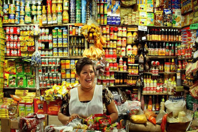 A Mexican middle-aged woman with a warm smile stands behind a counter laden with colorful packaged goods in her small, traditional Mexican grocery store. Shelves packed tightly with a wide variety of canned goods, bottles, and boxes rise behind her create a vibrant, somewhat cluttered backdrop. She wears a white apron dress over her sundress to protect it.
