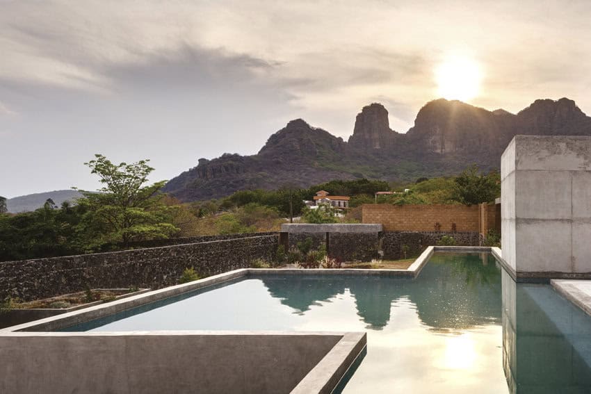 An outdoor photo of an infinity pool made with cantera stone. In the distance are a stone wall and a scrubby landscape with small mountains in the background.