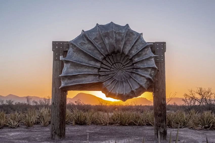 A large, weathered metal sculpture resembling an ammonite shell is mounted on two wooden posts, silhouetted against a vibrant sunset over a desert landscape. Mountains are visible in the distance, and sparse desert vegetation fills the foreground.