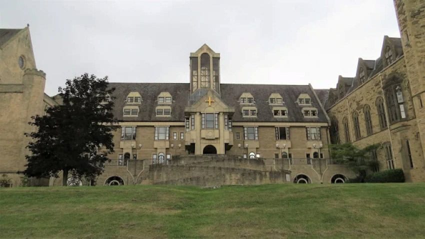 A hundreds of years old sandstone brick estate in Yorkshire with a small gold cross at the front entrance. At its front is an expanse of well-manicured lawn.