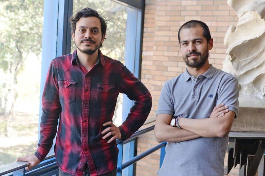 Two middle-aged Mexican male architects standing in a well-lit studio by a window, posing for a publicity photo.
