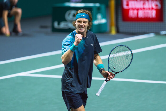 A male tennis player, Andrey Rublev, with a blue headband and wristbands clenches his fist and shouts in triumph on the court, holding his tennis racket in his other hand.