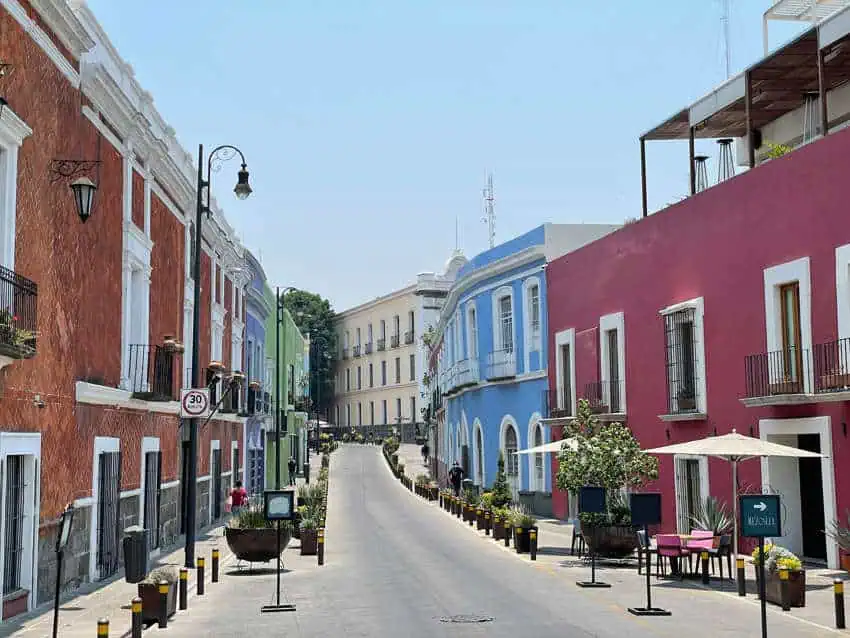 A sunny street in Puebla, Mexico, lined with historic buildings painted in vibrant colors like terracotta, blue, and hot pink, leading up a gentle incline.
