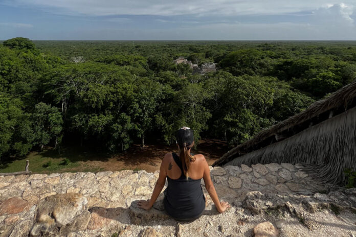 A person with their back to the camera sits on a stone edge, looking out over a vast, dense green jungle canopy that stretches to the horizon under a cloudy sky. In the distance, ruins are partially visible above the treetops.
