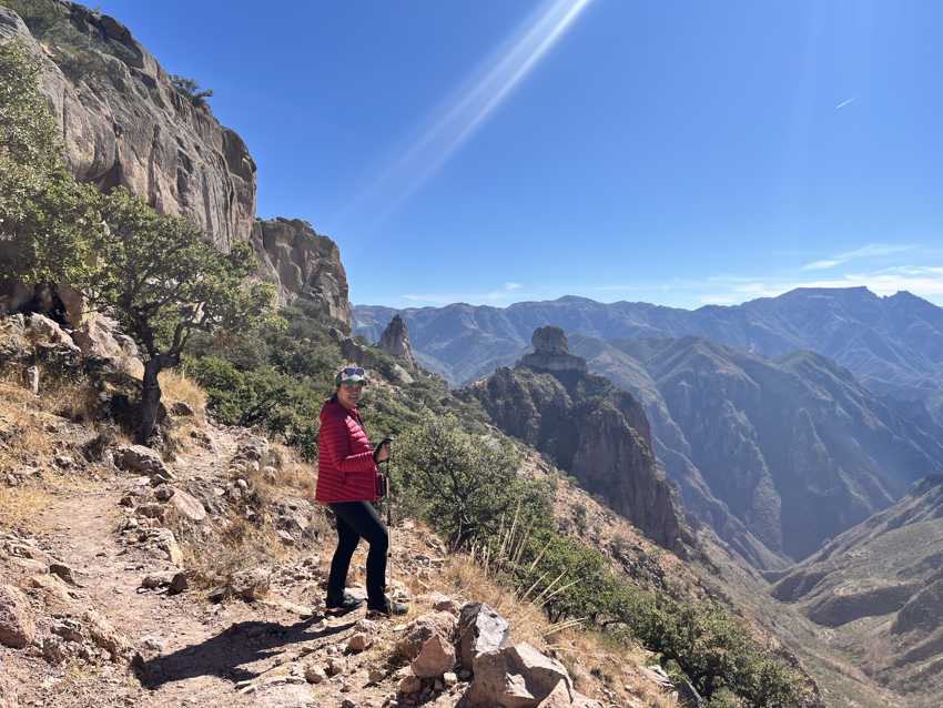 A hiker in a red jacket stands on a trail overlooking the vast, mountainous Copper Canyon in Chihuahua, Mexico