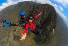 A woman in a red jacket smiles widely while tandem paragliding over a mountainous landscape with a professional pilot.