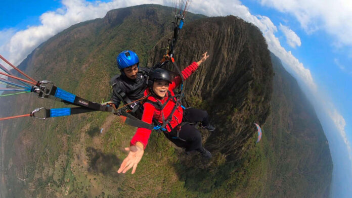 A woman in a red jacket smiles widely while tandem paragliding over a mountainous landscape with a professional pilot.
