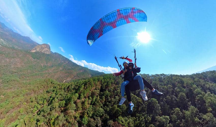 Two people tandem paragliding with a colorful canopy high above a lush green forest and mountains under a bright sunny sky in Mexico