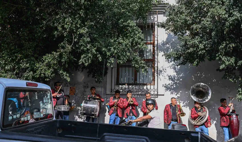 A group of male musicians in red jackets play various brass instruments and drums on a street, partially obscured by a parked truck and green trees, with a white building in the background.