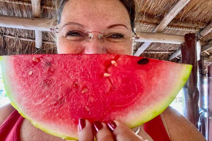 Woman posing in a Mexican palapa structure with a wedge of ripe watermelon in front of her face.