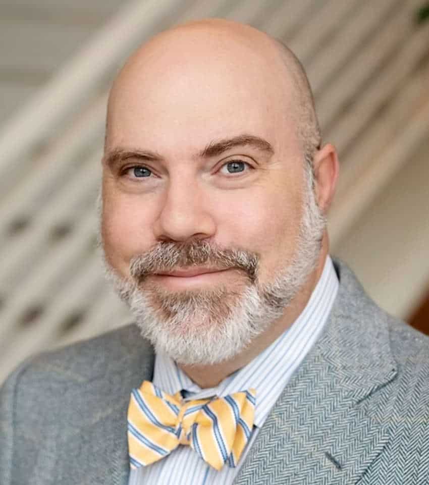 Headshot of a smiling bald man with a grey beard and blue eyes, wearing a grey textured blazer over a pinstriped shirt and a yellow and blue striped bow tie.