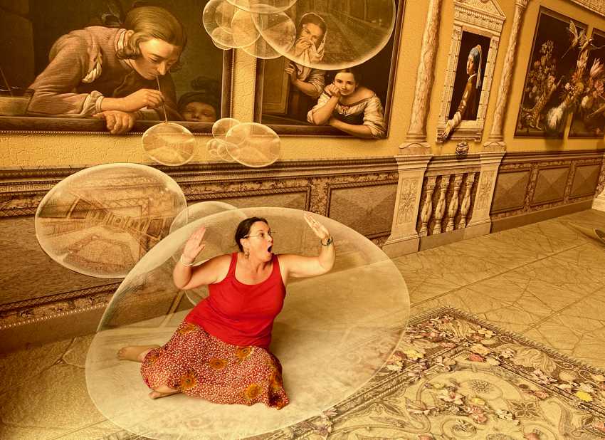 A woman in a red top and patterned skirt kneels on a rug, pretending to be trapped inside a large, transparent bubble, while surrounded by 3D optical illusion art depicting classic paintings with figures looking out from frames, and other bubbles floating around them.