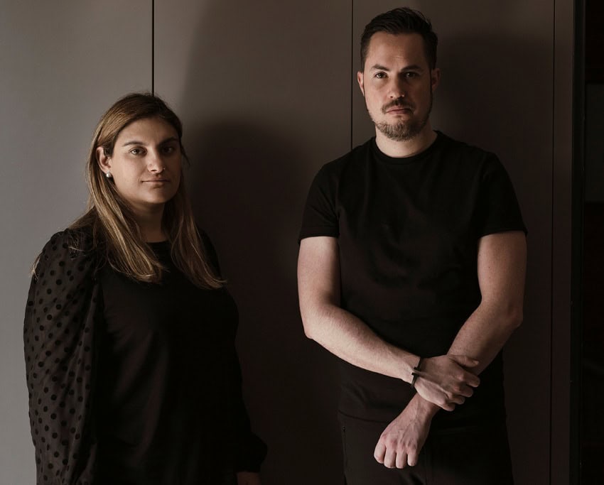 Mexican architects Marina Leboreiro and Carlos Faci posing against a dark wall for a publicity photo. They are both wearing black clothing and are in shadowy light.