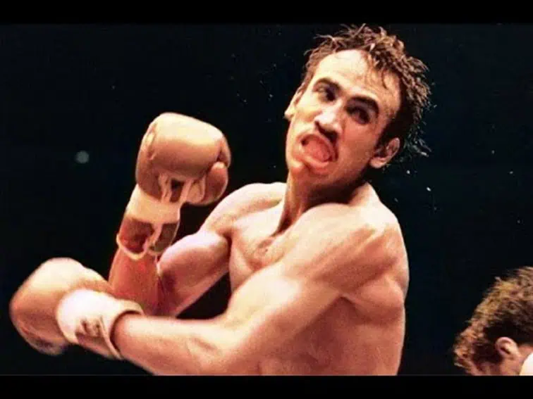 Mexican boxing champion Carlos Zarate in action, mid-fight. He has a mustache and short, dark, messy hair. His mouth is open, suggesting exertion or a shout, and his eyes are intensely focused. He is wearing light-colored boxing gloves and appears to be throwing or receiving a punch, with visible muscle tension in his arms, shoulders and face.