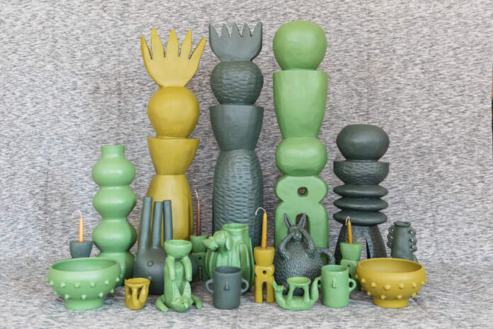 A collection of unique, handcrafted Mexican ceramic art pieces in shades of green and yellow, including tall stacked sculptures, vases, bowls, and whimsical figures, arranged on a textured grey surface.