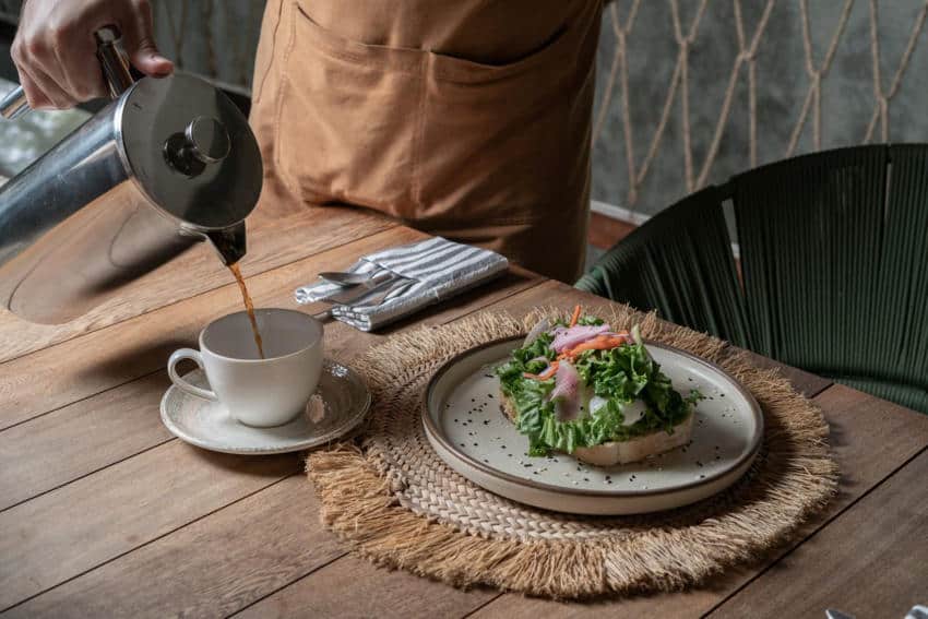 One of the locales in Bacalar's restaurant scene, Casa Hormiga. The photo shows a person pouring coffee into a cup next to a plate of avocado toast topped with greens and pickled vegetables on a rustic table in Mexico.