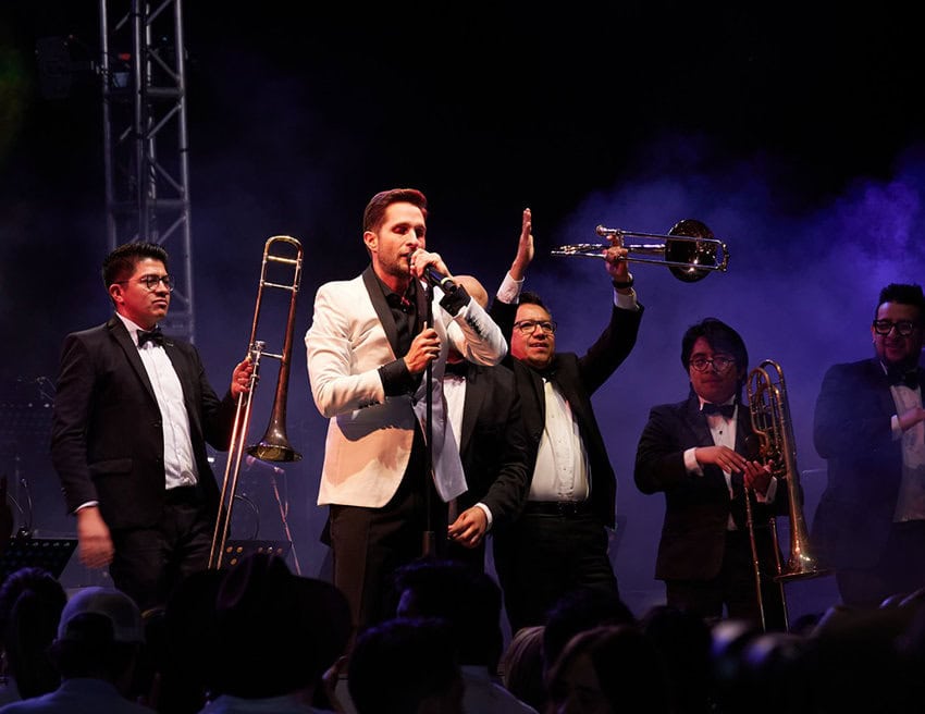 A male singer in a white jacket and black shirt holds a microphone, performing on stage with a band. Around him, four other male musicians in suits and bow ties play brass instruments, including a trombone and a trumpet, under dramatic purple stage lighting.