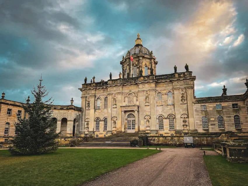The facade of a large English estate, Castle Howard, along with a manicured dirt road and a trimmed lawn leading to the estate.