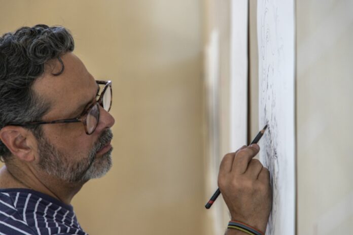 A man stands working on a pencil drawing mounted on a wall.