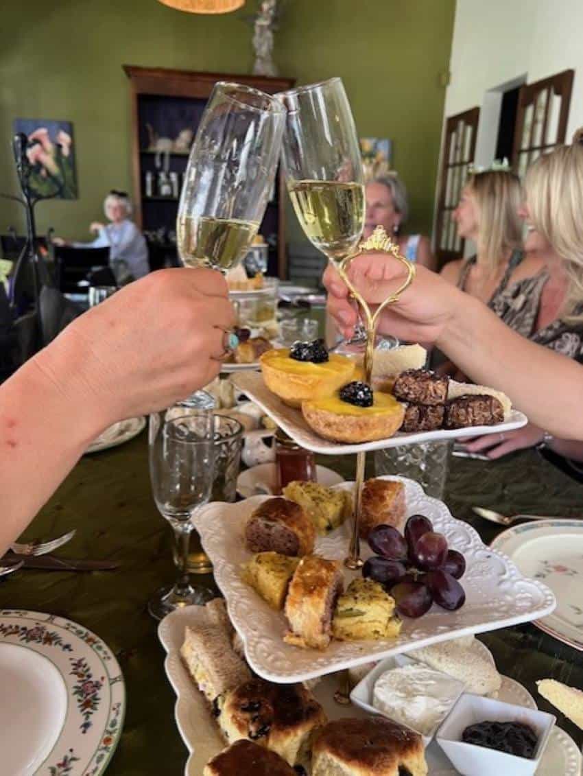 English teatime classic pastries and fruit on chinaware tiered serving trays filled to the brim. People sitting at the table are toasting to each other with filled champagne glasses.