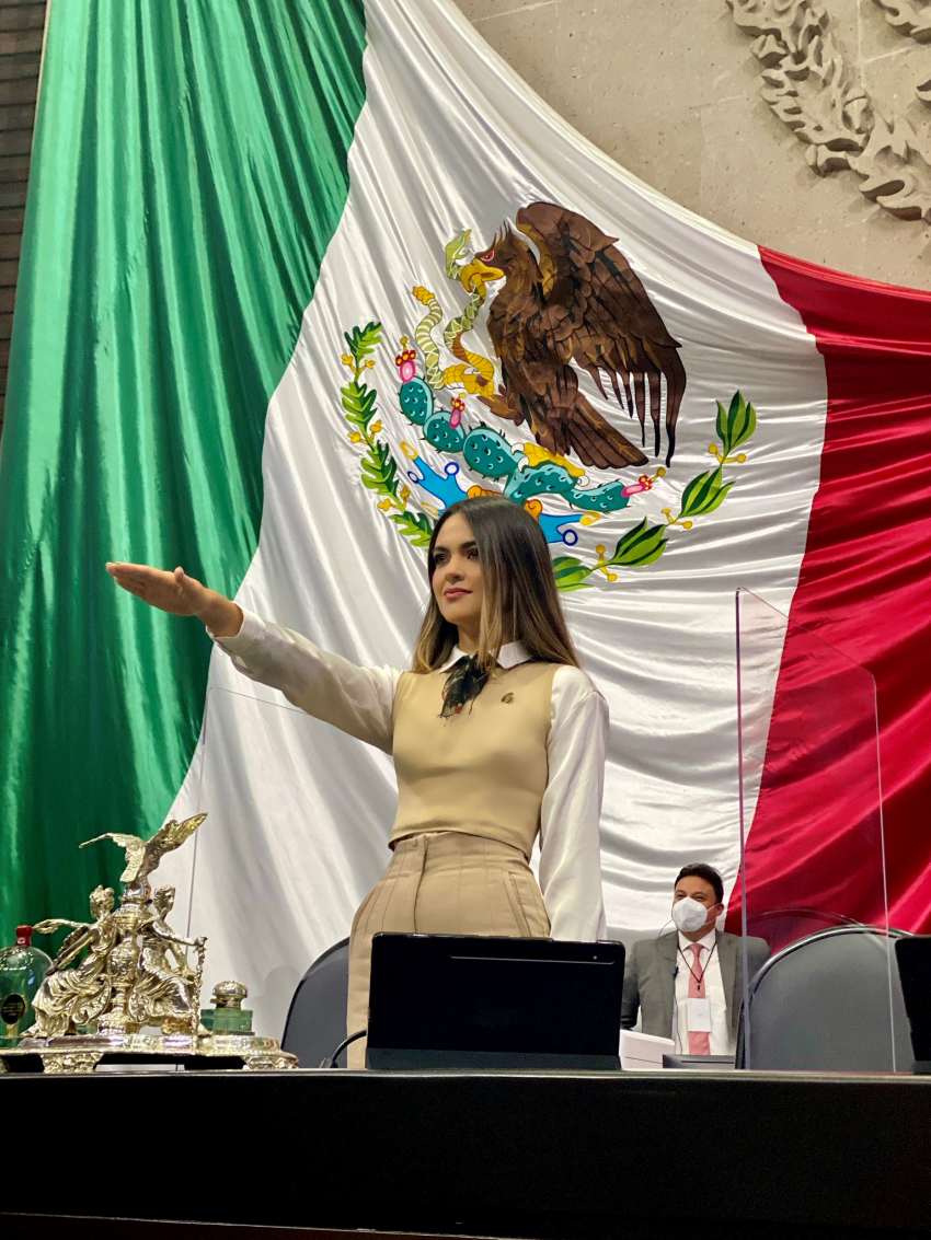 Current Chihuahua senator Andrea Chavez Trevino being sworn in in 2021 as a federal deputy. She has her arm extended forward in front of her as she takes the oath of office. Behind her is a massive Mexican flag on the legislature wall.