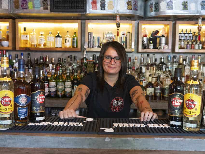 A smiling bartender with tattooed arms and glasses, Claudia Cabrera, stands behind a bar filled with bottles of liquor.