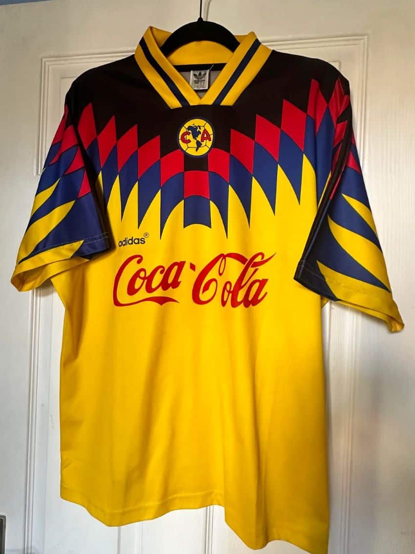 A yellow soccer team uniform jersey for Club America soccer team in Mexico City, on a hanger hanging from a interior white door in a residential home. It has and navy blue diamonds laid out in a zigzag pattern, the Club America logo just under the V-neck, and the Coca Cola and Adidas trademarked names toward the jersey's center