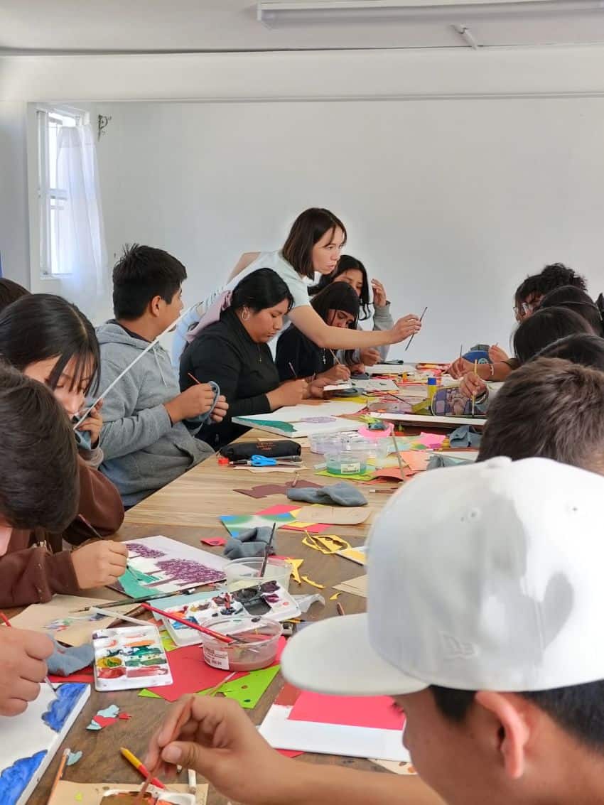 An art teacher demonstratively holds out a artistic paintbrush toward a student across a large table where many other adult students sit working on painting projects.