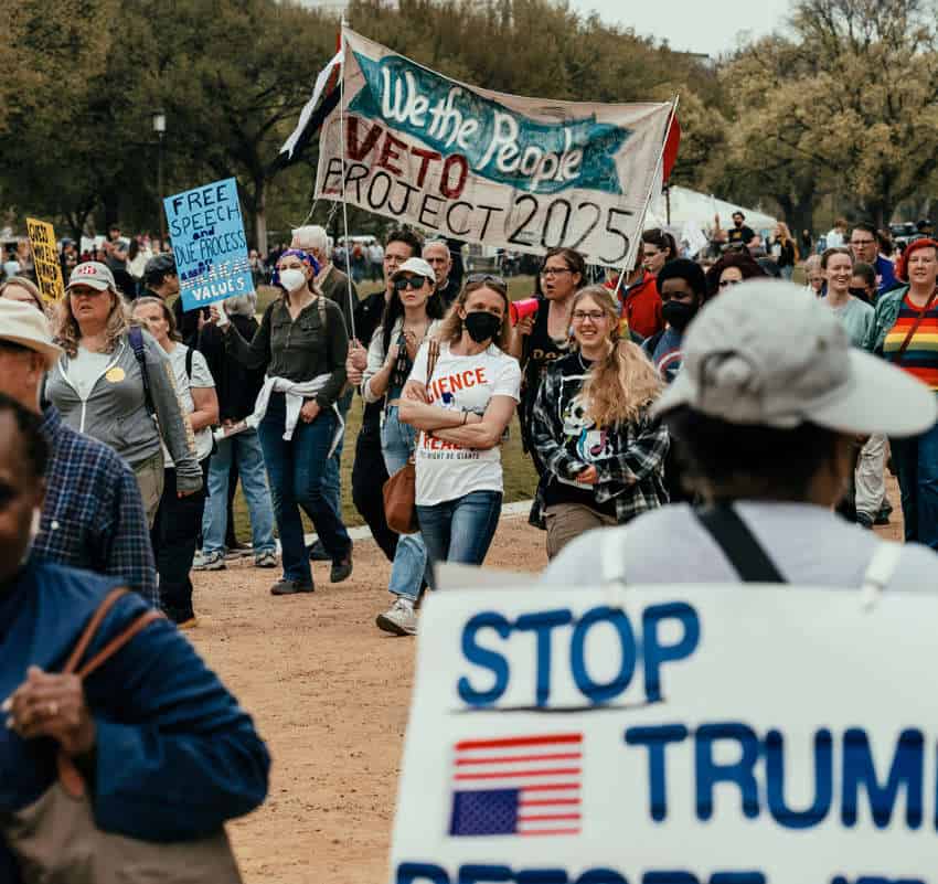 A crowd of people protesting and marching, two people carrying a sign saying "We the People veto Project 2025." There are other signs regarding free speech and due process and stopping Donald Trump