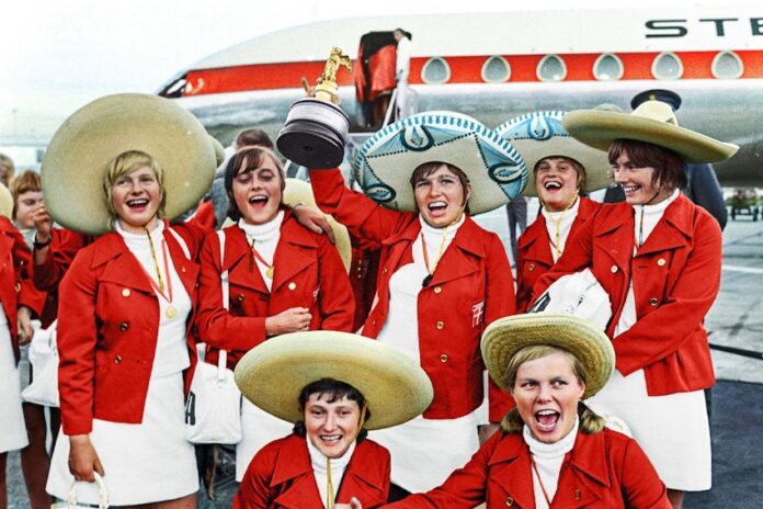 A team of women in Red jackets and sombreros lift the world cup trophy on the steps on a plane.