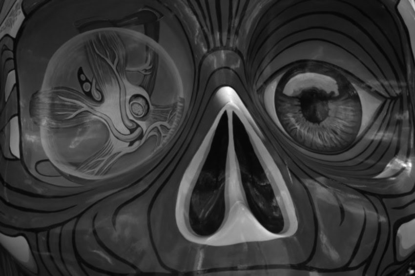 A black and white close-up of a skull-like face. On the right, a detailed human eye is depicted, while on the left, an abstract, almost biological or neurological pattern is visible within a rounded shape, suggesting a cross-section or inner workings. The face has prominent nose holes and stylized lines radiating outwards.