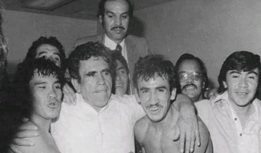 A vintage black-and-white photograph of a group of boxers and boxing associates surrounding Arturo "Cuyo" Hernandez, a Mexican boxing manager and promoter. All the men are gathered closely together, many with their arms around each other. In the center, a man with curly hair and a white shirt has his arms around two younger shirtless menAnother man in a suit and mustache stands taller behind the group, looking over their heads.