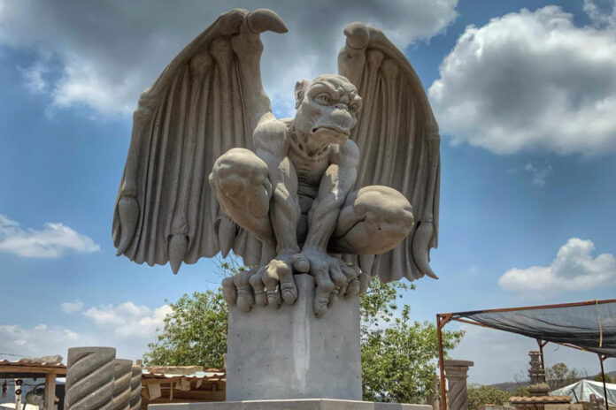 A large, light-colored cantera stone sculpture of a crouching gargoyle with outstretched wings, set against a blue sky with some clouds.