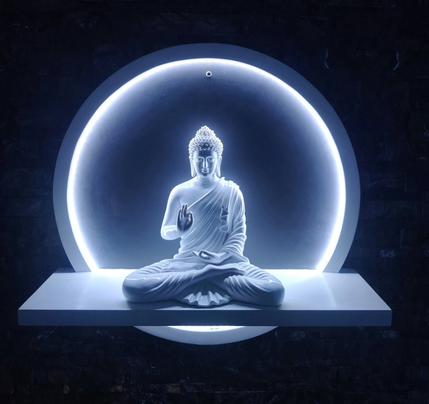 A white statue of Buddha in a meditative pose, seated on a white shelf. Behind the statue, a bright white LED ring light creates a halo effect, illuminating the figure against a dark, textured background.