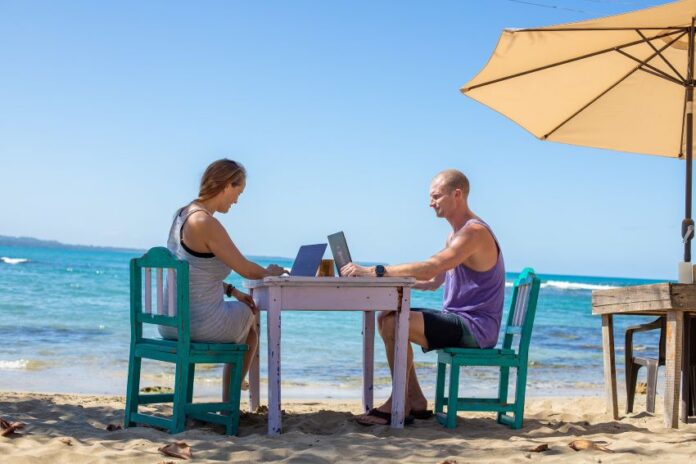 Man and woman work on laptops at a table on a beach